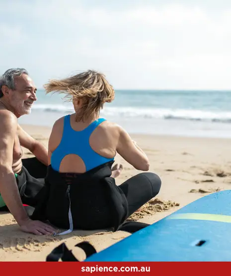 elder man and woman sitting on sand wearing wetsuits while having a conversation